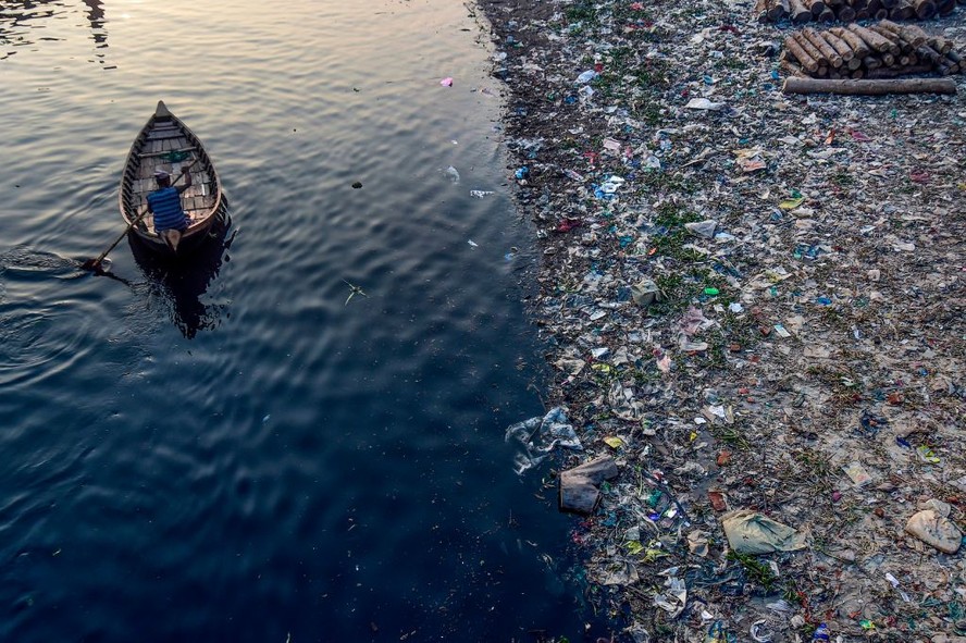 Homem rema em um barco enquanto plásticos flutuam na superfície do rio Buriganga, em Bangladesh