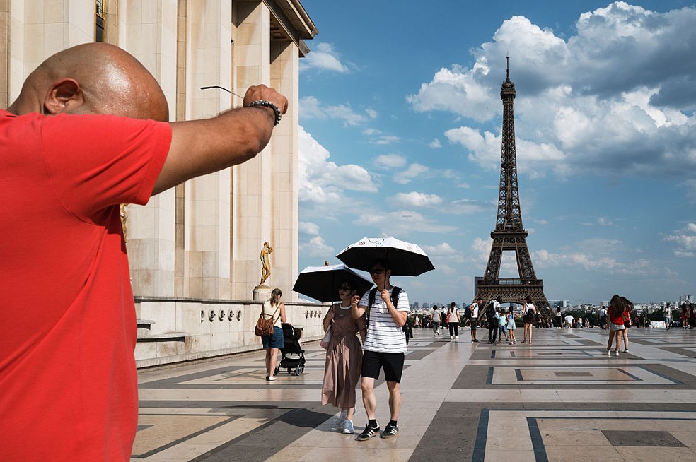 Homem sua sob calor intenso enquanto turistas usam guarda-chuva para se proteger do sol na região do Trocadéro, em frente à Torre Eiffel, durante onda de calor na França, em junho de 2026. — Foto: Jerome Gilles/NurPhoto via Getty Images