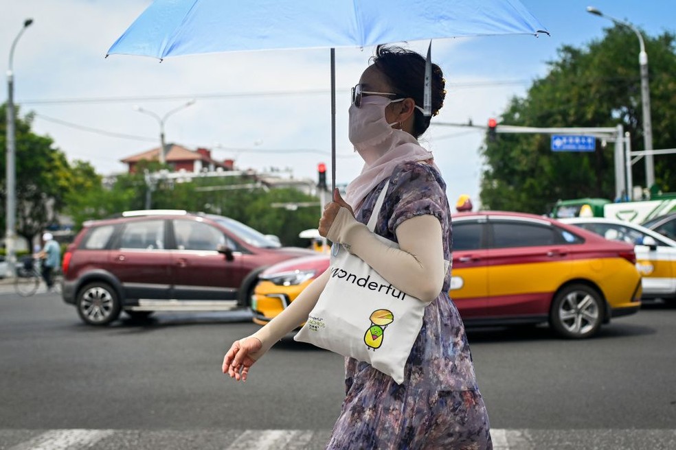 Uma mulher usando um guarda-chuva e roupas de proteção contra o sol atravessa uma rua em Pequim, em julho de 2023 — Foto: Getty Images